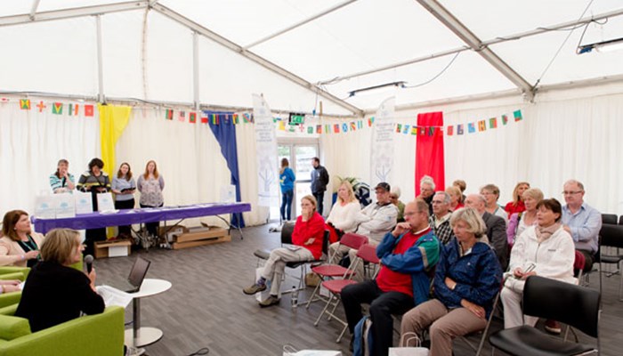 A photo of the crowd of adults sitting on red fold out chairs at a Family History Q&A, with panel members at the front and a stall of leaflets and goody bags in the background.