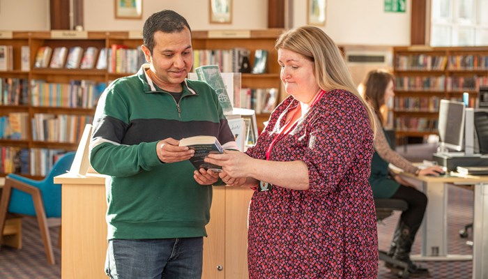 A member of staff showing a customer a book in Knightswood Library. There are library shelves in the background and someone sitting at a computer.
