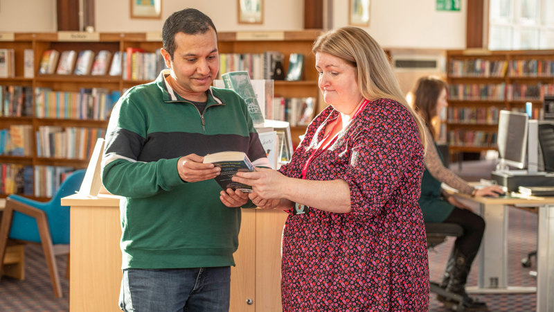A member of staff showing a customer a book in Knightswood Library. There are library shelves in the background and someone sitting at a computer.