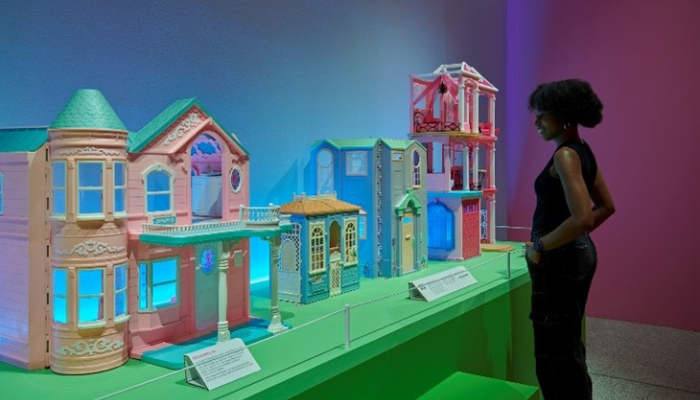 A visitor stands in front of a glowing display of Barbie dream houses in pastel pink, blue, and purple, arranged in a row to resemble a colourful street.