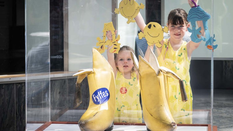Two children holding colourful crafts behind a display case with large banana-shaped boots.