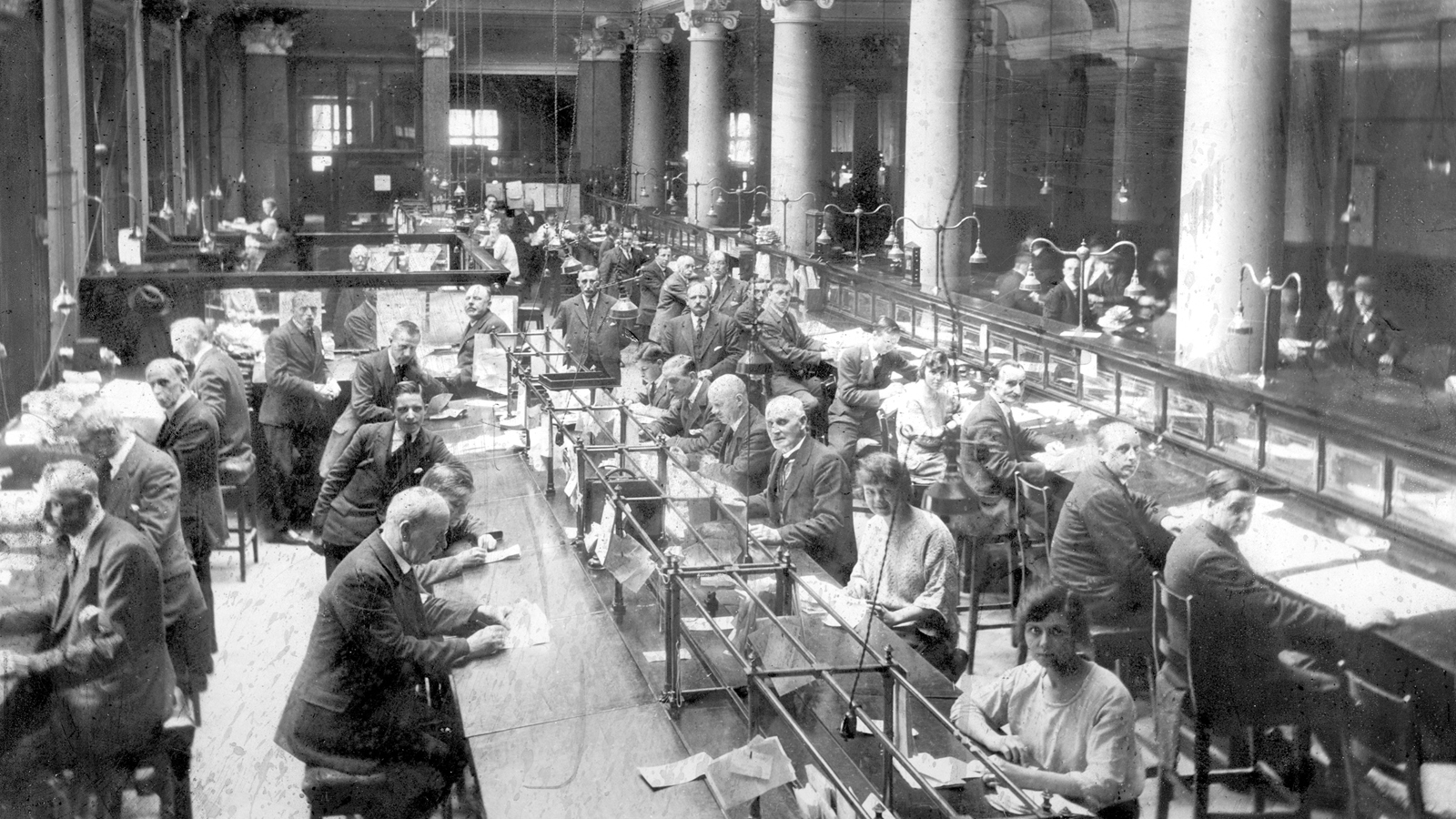 A black and white photograph of workers in office attire, sitting at designated tables covered in papers, working in a large room held up with many architectural pillars.