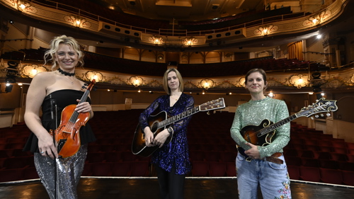 Three musicians standing on stage at a theatre holding their instruments
