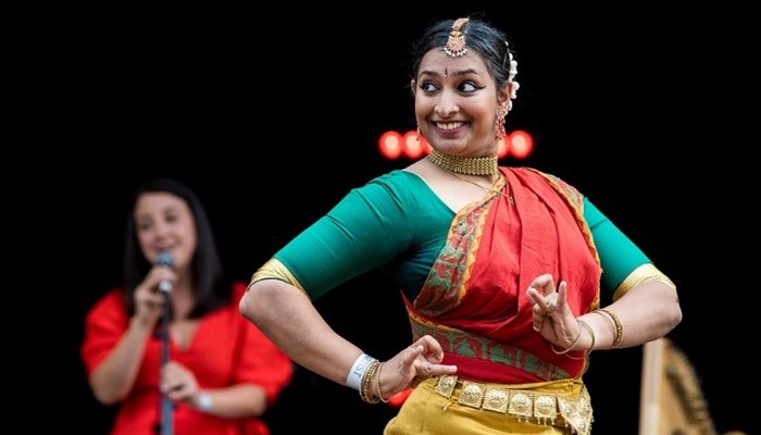 A person in colourful dress and head piece is smiling and performing on a stage. There is a person singing into a microphone behind them.