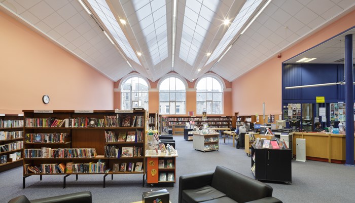 Main view of the library interior which had high ceilings and large windows. The roof is a white saw-style which is also a skylight. The walls are peach and blue and there are dark walnut bookshelves around the perimeter and middle of the room. There are 3 large arched windows at the end of the room. There is also a coffee table surrounded by soft black leather chairs. 