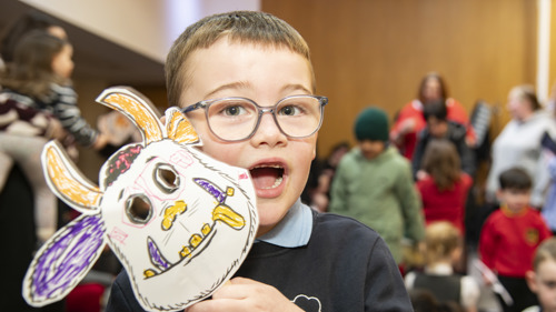 A young person wearing glasses is taking part in a group craft event in a library. They are holding up a colourful paper mask of the Gruffalo character