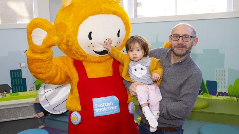 Bookbug in Partick Library. Adult holding a small child who is squeezing the Bookbug mascot's nose. Bookbug is a yellow smiling bug developed by the Scottish Book Trust. They have wings and antennae and wear red dungarees. 