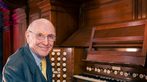 Bill sits at the organ in Kelvingrove Museum smiling at the camera, he is wearing glasses and a grey suit