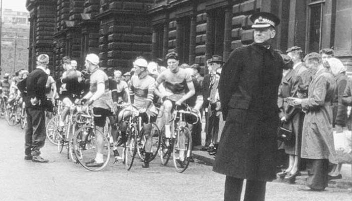A black and white photo of a queue of people lining up on their bikes and security at the forefront.