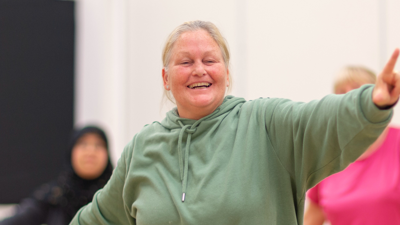 A person smiling during a wellbeing class with their left hand in the air pointing