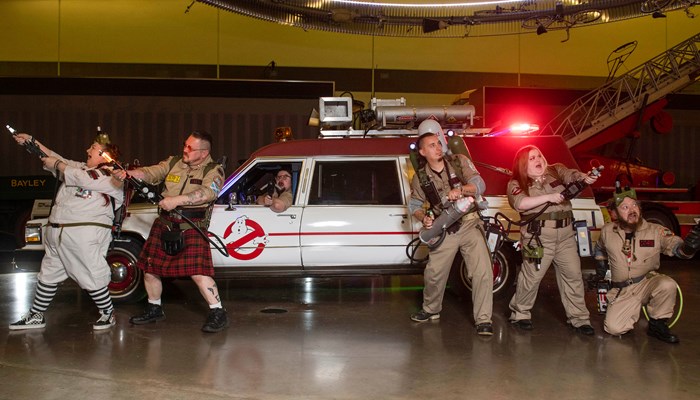 Five people dressed in jumpsuits standing in front of the car from the Ghostbusters film another person sitting inside the car