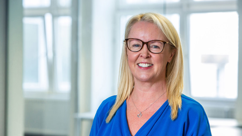 A person with long blonde hair, wearing dark rimmed glasses and a blue top, smiling in front of large windows in an open plan office space