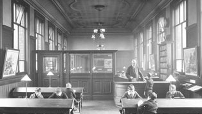 Black and white image showing an old fashioned library, there are large wooden desks and people sitted at them reading books and newspapers