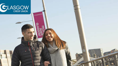 Two people walking arm in arm along a city centre river on a clear winter day. There are flags behind them that read 'People Make Glasgow'