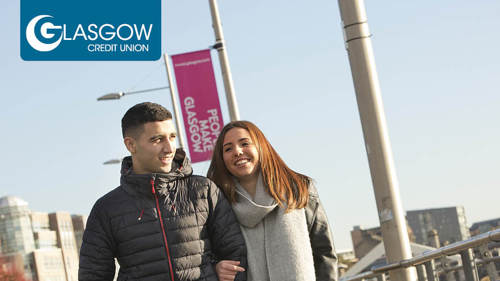 Two people walking arm in arm along a city centre river on a clear winter day. There are flags behind them that read 'People Make Glasgow'
