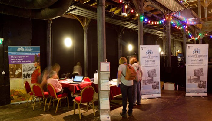 A photo of staff setting up a stall at an event with a red table cover, Family History pop up banners, laptops, fairy lights, leaflets and goody bags.