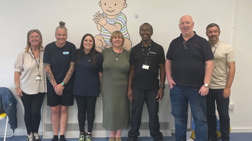 A group of seven people are standing in a line for a photograph in a community hall with a large mural of a cartoon baby painted on the wall behind them