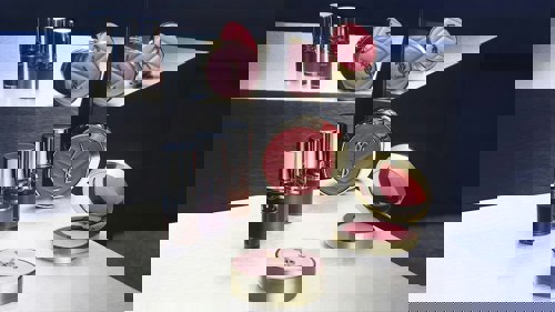 An arrangement of luxury Yves Saint Laurent makeup products displayed on reflective metallic surfaces, including round gold-and-pink compact cases with the YSL logo and several lip gloss or liquid lipstick tubes in various shades, all positioned near a mirrored backdrop