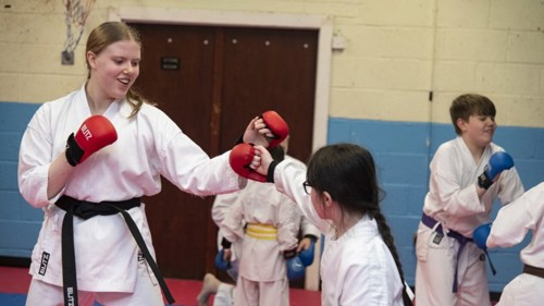 An adult instructor is taking a Karate class for young people. They are all wearing white Karate uniforms and wearing small punch gloves to practice