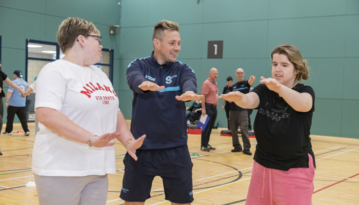 Three adults smiling and exercising with outstretched arms in a large, brightly lit indoor sports hall.