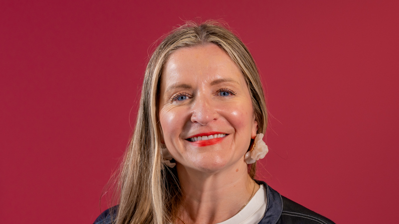 A woman with light brown hair smiles in front of a rubine background
