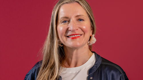 A woman with light brown hair smiles in front of a rubine background