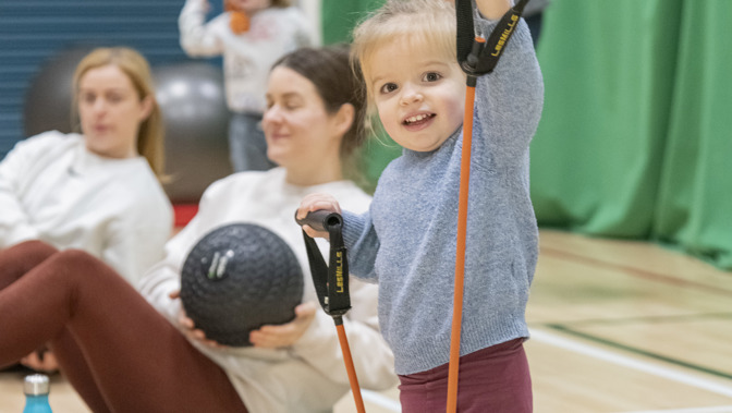 A toddler stands on a gym mat in a sports hall, smiling while holding orange resistance bands raised in both hands. The child wears a blue sweater, purple leggings, and white socks. Behind them, adults sit on mats with exercise equipment, and green wall padding and gym flooring are visible in the background.