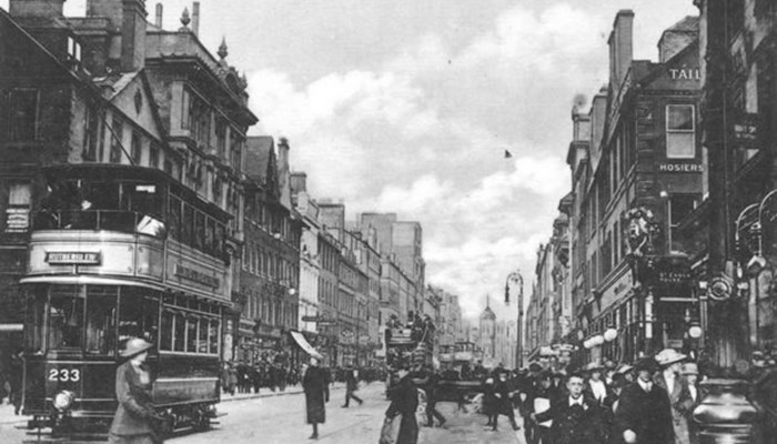 A black and white photo of a busy street with trams, pedestrians and many sandstone tall shop buildings with clouds in the sky