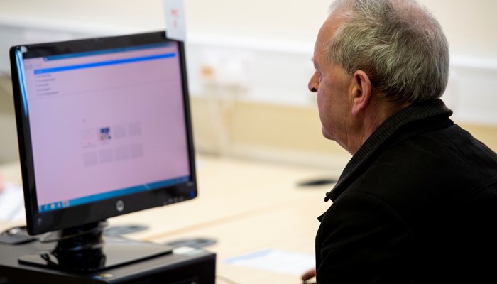 An adult sits at a PC. They have grey hair and a black jacket.