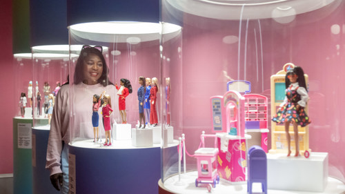 A person is smiling and looking at several Barbie dolls in a glass tube display in a pink exhibition room