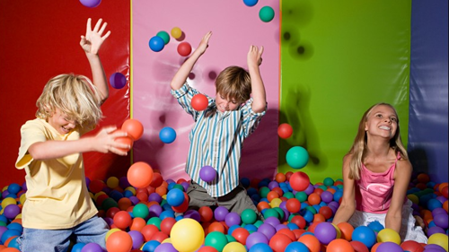 Children in a ball pit at a soft play smiling and throwing balls in the air against a striped colourful background