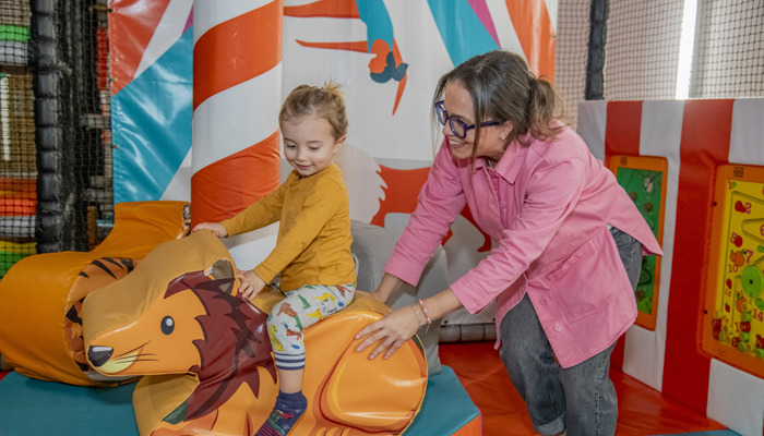 A parent pushing a young child on a soft play lion, both of them are smiling.