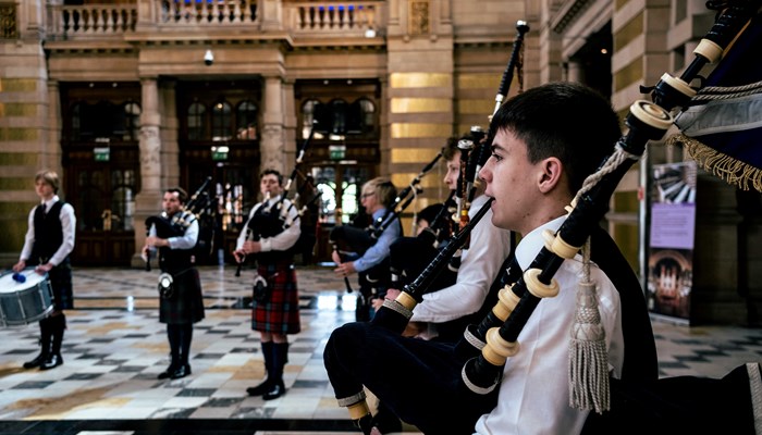 Five smartly dressed young bagpipers and drummers play together in a grand Victorian setting, the main hall of Kelvingrove art gallery and museum