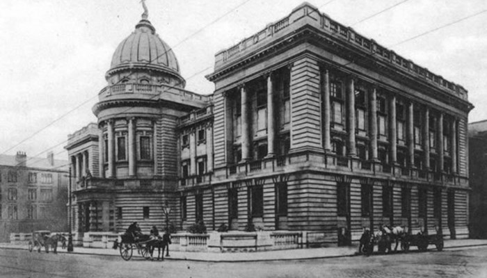 A black and white photo of the side of The Mitchell Library, home of The Family History centre. Horse and carriages are transporting people around the streets at either side and there are tenement houses in the background.