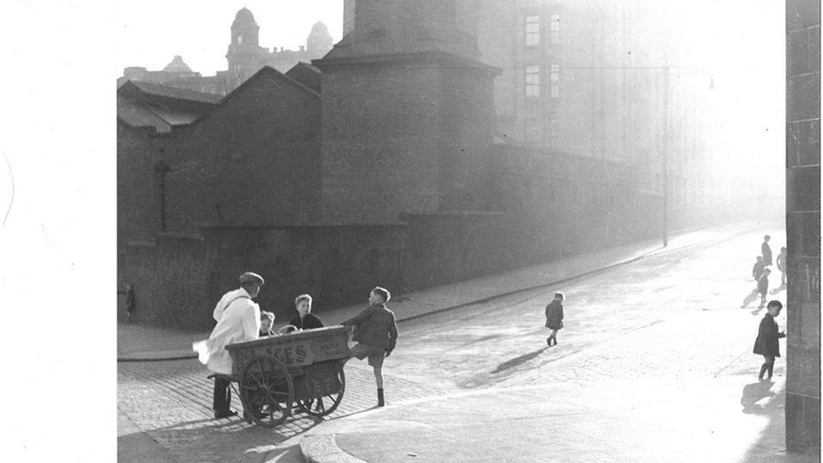 A black and white photograph of young kids playing on sunny cobbled street, some of which are chatting to an adult who is pulling along a small cart.