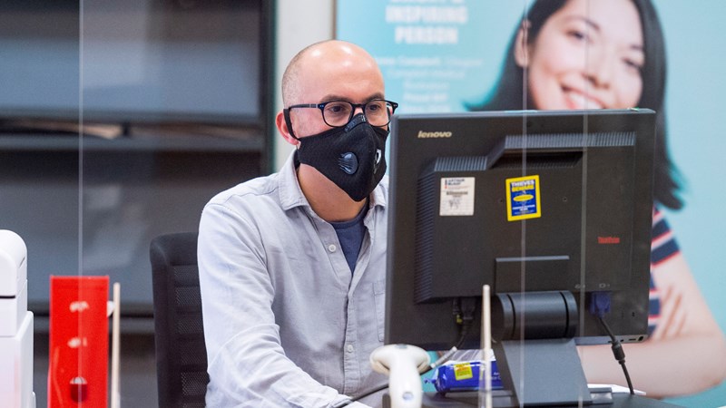 A man wearing a shirt and a face mask sitting in front of a PC in an office setting